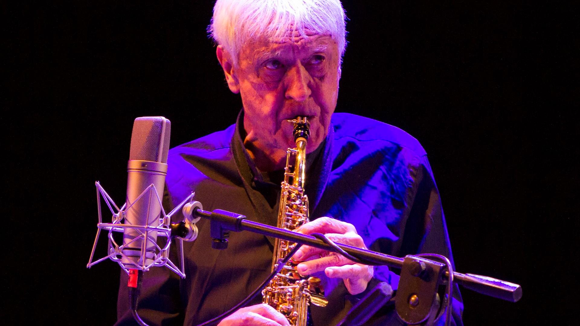 Portal steht auf der Bühne, bläst in sein Instrument und blickt nach vorne. Er wird blau angestraht, der Hintergrund ist dunkel. 