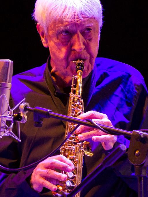 Portal steht auf der Bühen, bläst in sein Instrument und blickt nach vorne. Er wird blau angestraht, der Hintergrund ist dunkel. 