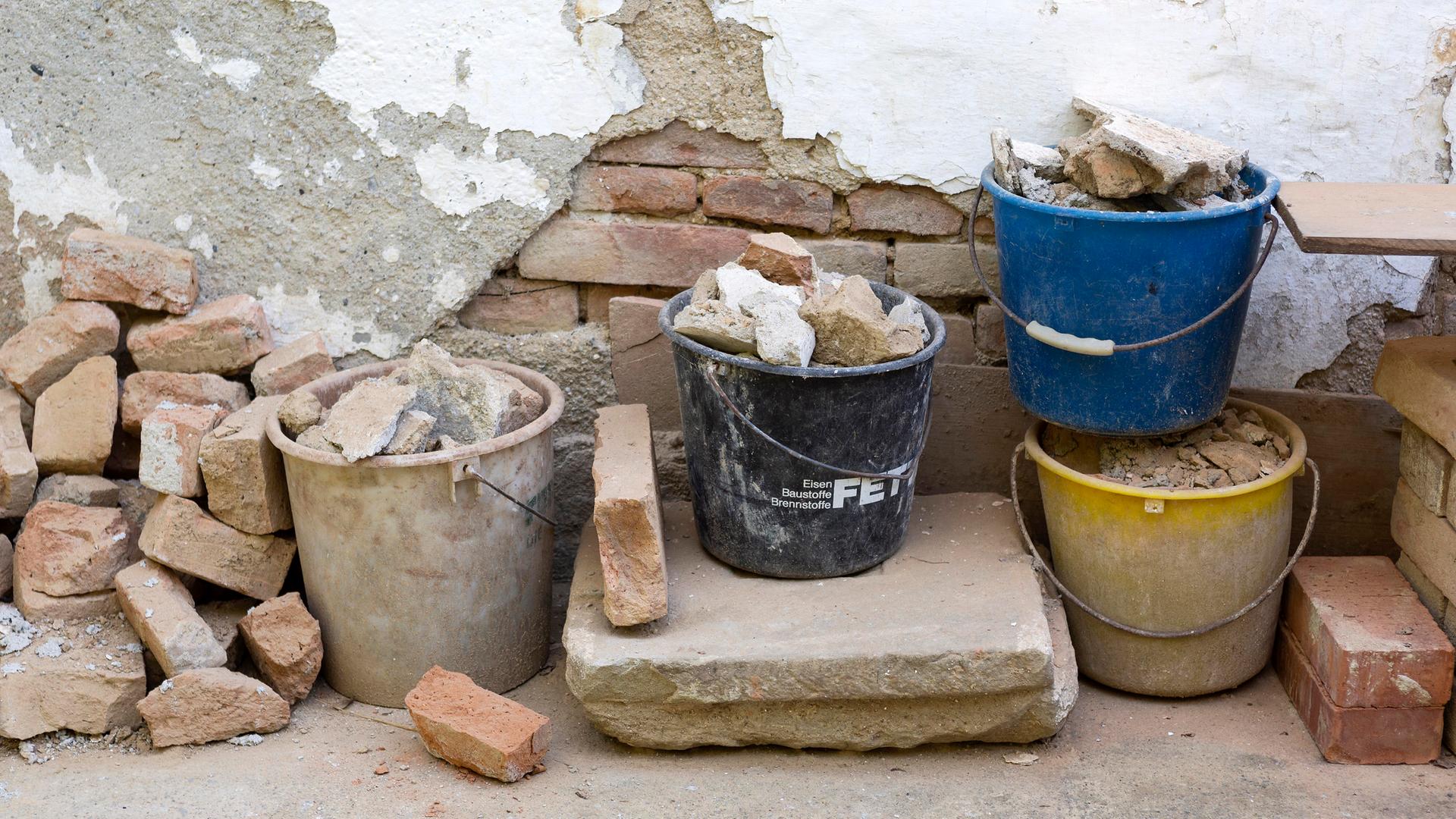 Wand eines Altbaus mit abgebröckeltem Putz, darunter Ziegelmauerwerk, davor Eimer mit losen alten Ziegeln