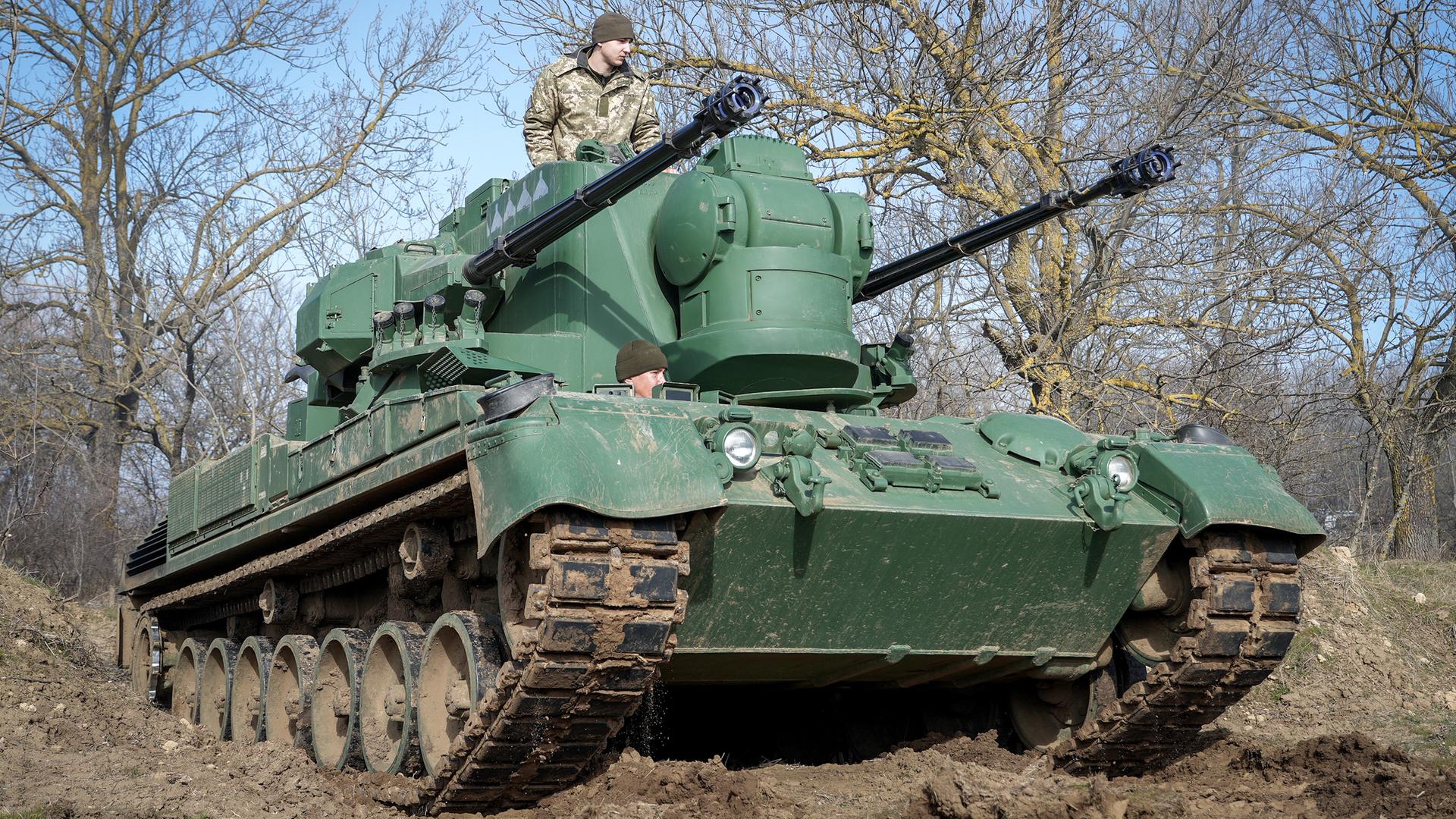 Ein Panzer mit großen Kanonen-Rohren fährt an Bäumen vorbei.