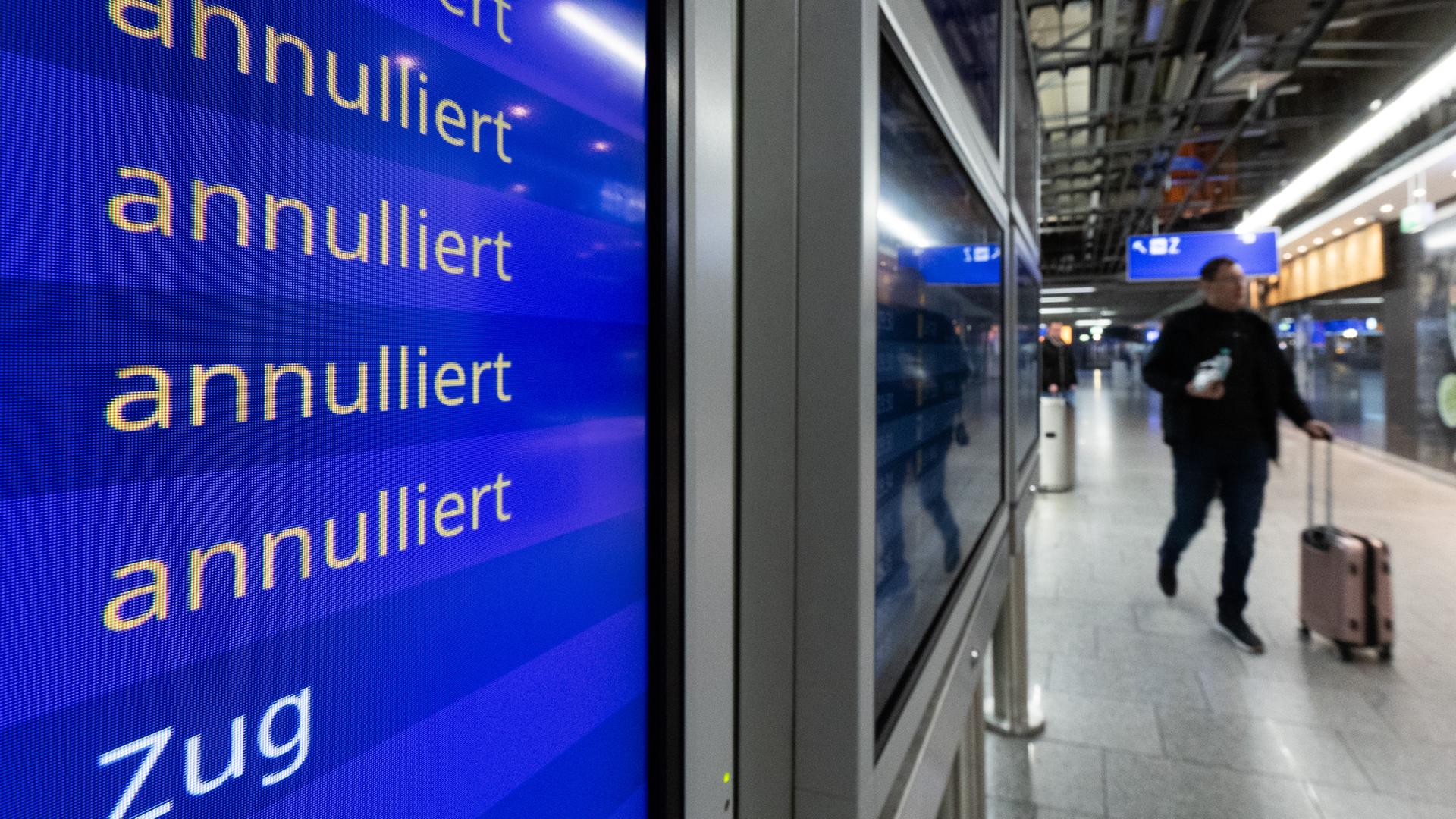 Eine Anzeige in einer Abflughalle weist mehrere Flüge als annulliert aus. Im Hintergrund ein Reisender mit Rollkoffer.