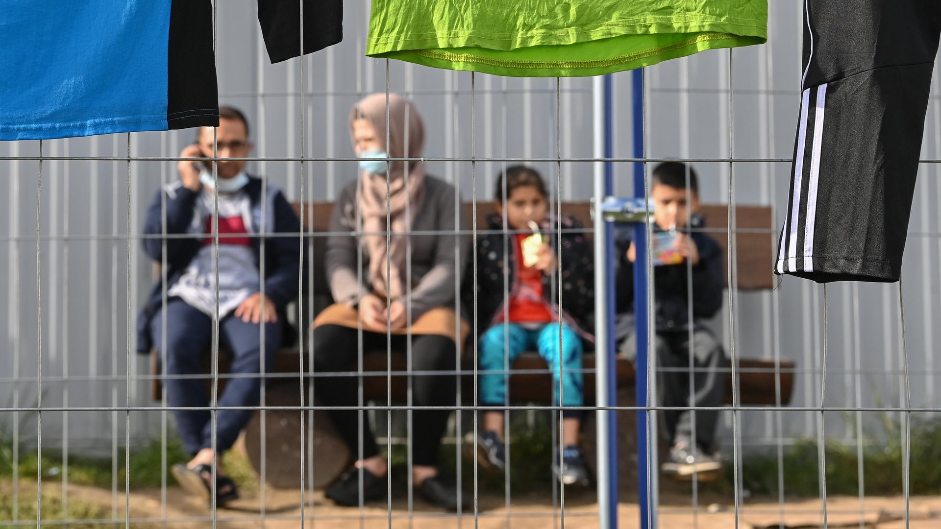 Ein Mann, eine Frau un zwei Kinder sitzen auf einer Bank an der Wand eines Containers hinter einem Zaun.