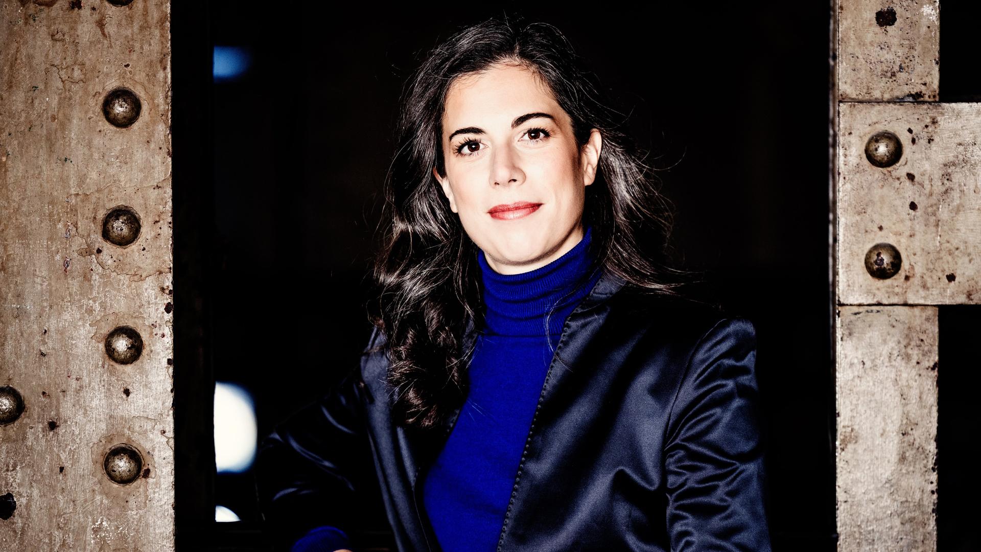 Eine Frau mit dunklen, längeren Locken steht zwischen industriell wirkenden Metallstreben und blickt offen in die Kamera. Sie trägt einen knallblauen Rolli und ein dunkelblau glänzendes Jackett. 
