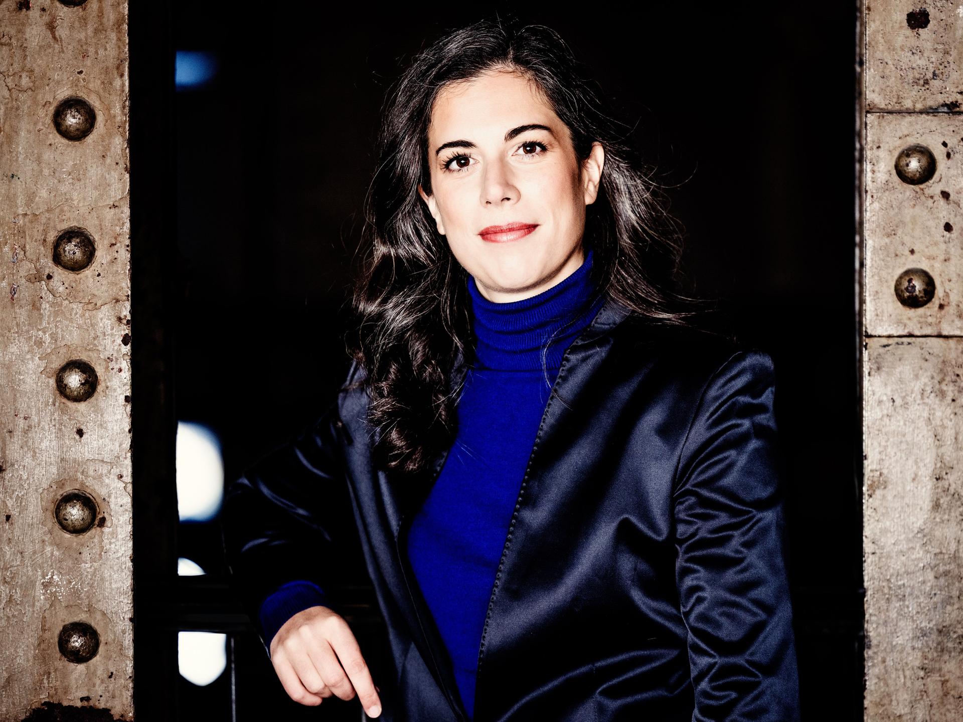 Eine Frau mit dunklen, längeren Locken steht zwischen industriell wirkenden Metallstreben und blickt offen in die Kamera. Sie trägt einen knallblauen Rolli und ein dunkelblau glänzendes Jackett. 