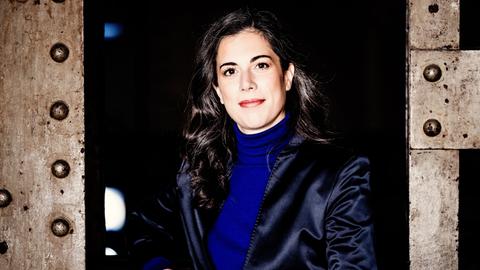 Eine Frau mit dunklen, längeren Locken steht zwischen industriell wirkenden Metallstreben und blickt offen in die Kamera. Sie trägt einen knallblauen Rolli und ein dunkelblau glänzendes Jackett. 