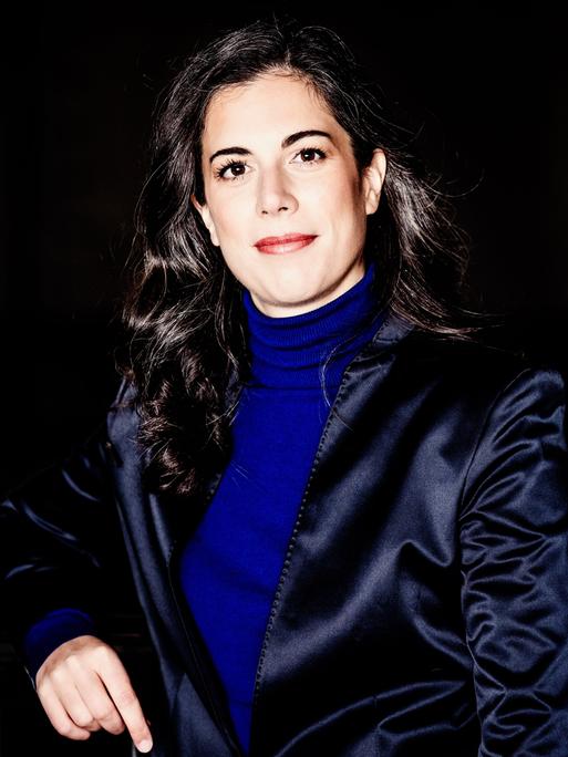 Eine Frau mit dunklen, längeren Locken steht zwischen industriell wirkenden Metallstreben und blickt offen in die Kamera. Sie trägt einen knallblauen Rolli und ein dunkelblau glänzendes Jackett. 