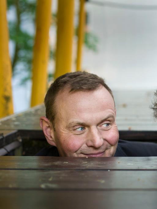 Devid Striesow und Axel Ranisch schauen aus einer hölzernen Bodenluke hervor und schauen sich verschmitzt an