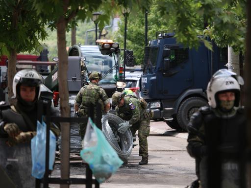 Zwischen  lokalen Polizeikräften sind KFOR Soldaten in Tarnuniformen zu sehen, die Natodraht ausrollen.
