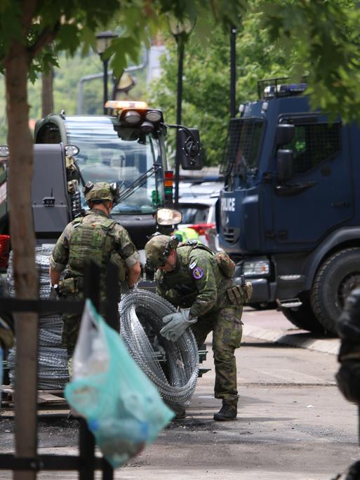 Zwischen  lokalen Polizeikräften sind KFOR Soldaten in Tarnuniformen zu sehen, die Natodraht ausrollen.