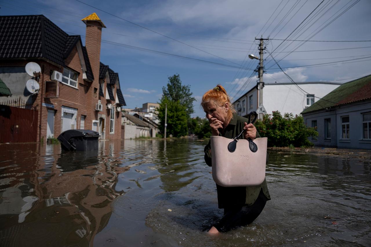 Eine Frau watet durch knietiefes Wasser, im Hintergrund sind Häuser und Bäume zu sehen.