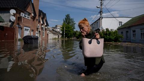 Eine Frau watet durch knietiefes Wasser, im Hintergrund sind Häuser und Bäume zu sehen.