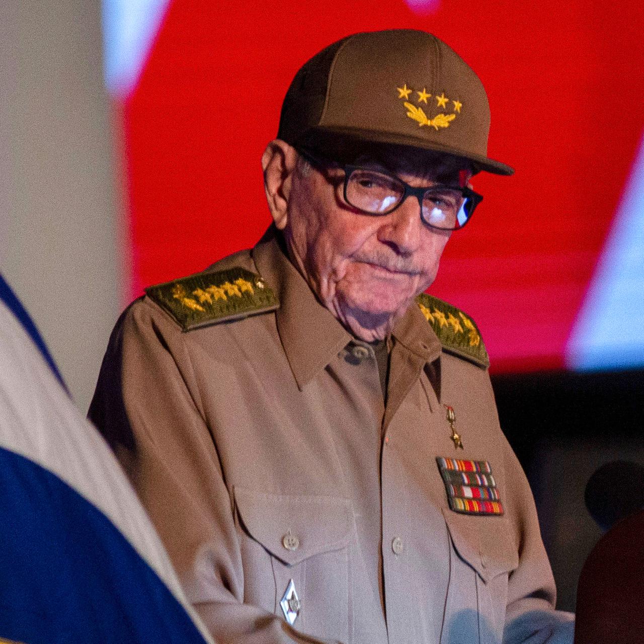 Ein alter Mann mit Brille und in Uniform - es ist Raúl Castro. Er sitzt neben einer kubanischen Flagge.