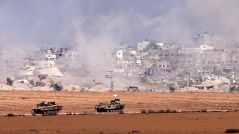 Panzer fahren an der Grenze zum Gazastreifen entlang. Im Hintergrund sind Gebäude einer Ortschaft und Rauch zu sehen.