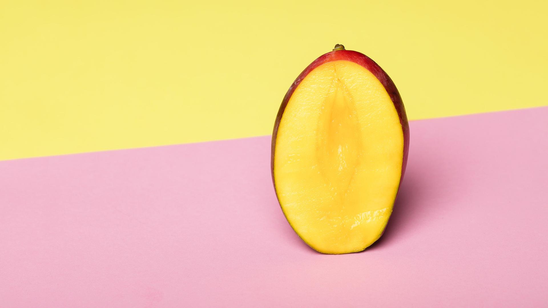 Eine aufgeschnittene Mango steht vor einem gelben Hintergrund auf einer rosa Fläche. 