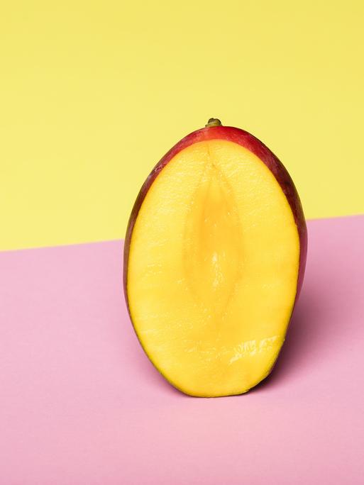 Eine aufgeschnittene Mango steht vor einem gelben Hintergrund auf einer rosa Fläche. 