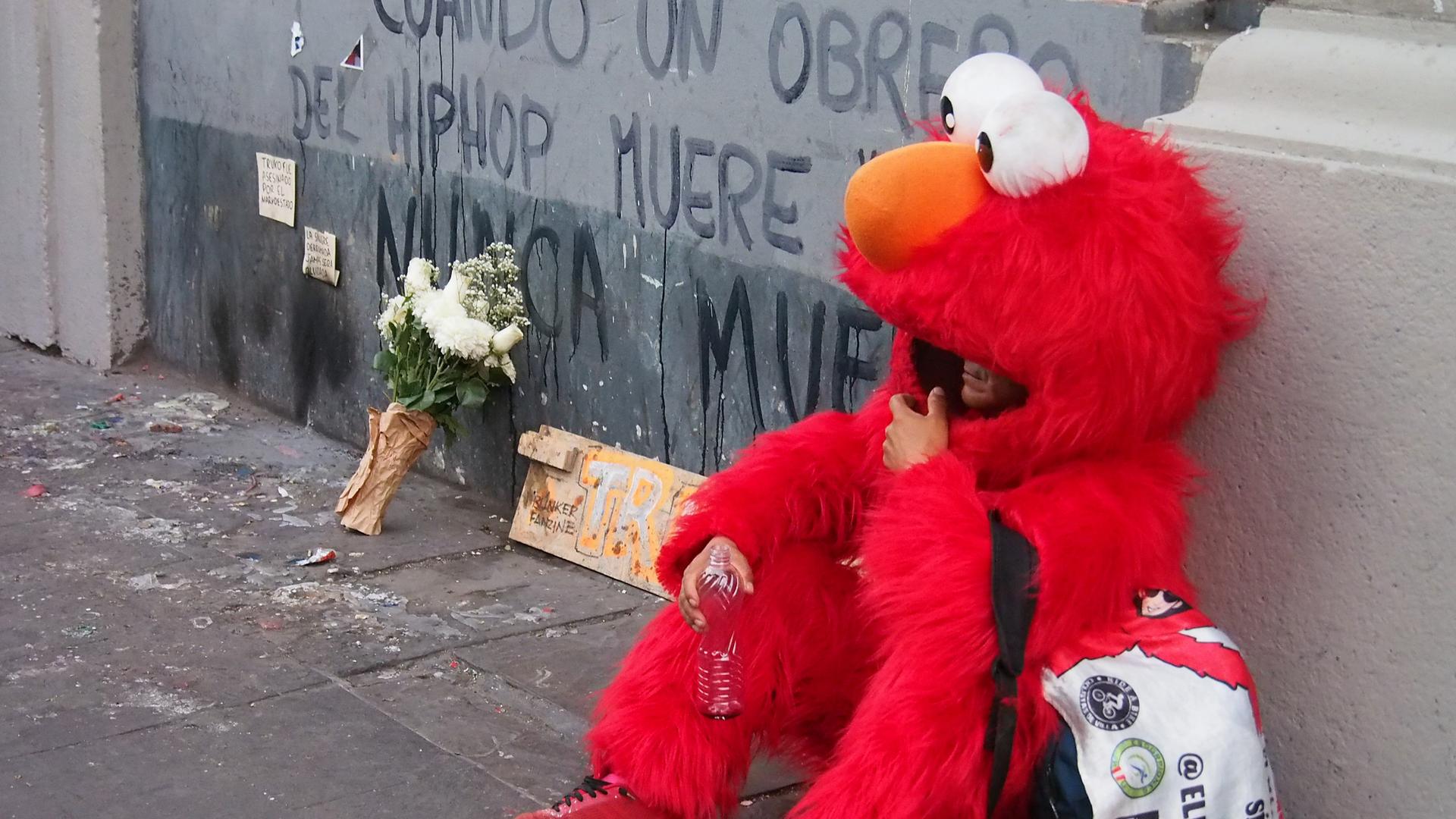 „Wenn ein Hip-Hop-Künstler stirbt, stirbt er nie“ – steht an einer Wand. Davor sitzt eine Person, die als Elmo aus der Sesamstrasse verkleidet ist.