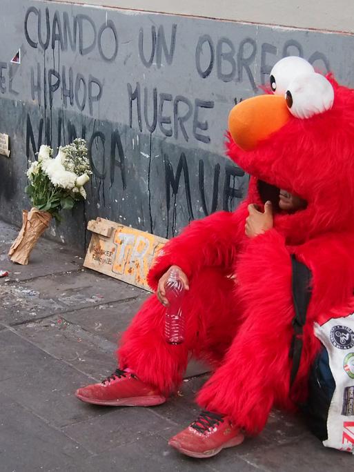 „Wenn ein Hip-Hop-Künstler stirbt, stirbt er nie“ – steht an einer Wand. Davor sitzt eine Person, die als Elmo aus der Sesamstrasse verkleidet ist.