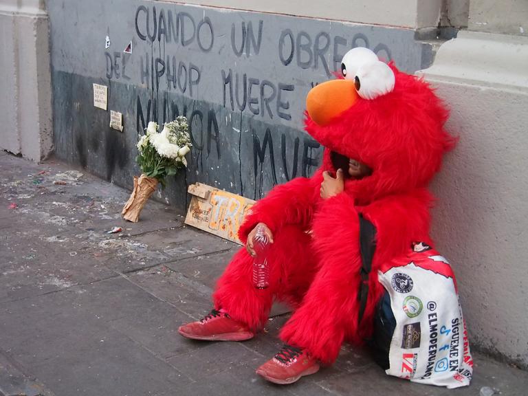 „Wenn ein Hip-Hop-Künstler stirbt, stirbt er nie“ – steht an einer Wand. Davor sitzt eine Person, die als Elmo aus der Sesamstrasse verkleidet ist.