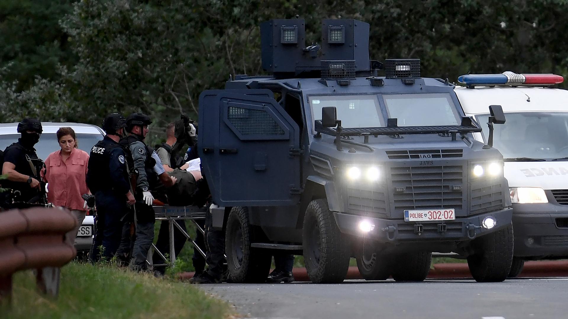 Im Schutz eines gepanzerten Polizeifahrzeugs laufen mehrere Polizisten in Kampfmontur und ein Zivilist. Daneben weitere Autos.