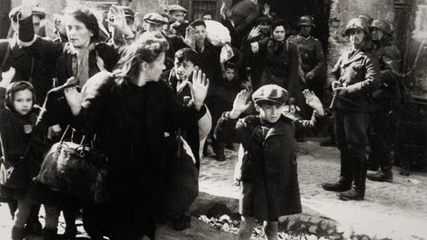 Warschauer Ghettoaufstand April/Mai 1943: Kleiner Bub mit erhobenen Händen, Frauen und andere Kinder kommen aus einem von den deutschen Truppen eroberten Haus. Aus dem Album von SS-Gruppenführer und Polizei-Generalmajor Jürgen Stroop. Photographie, 1943.