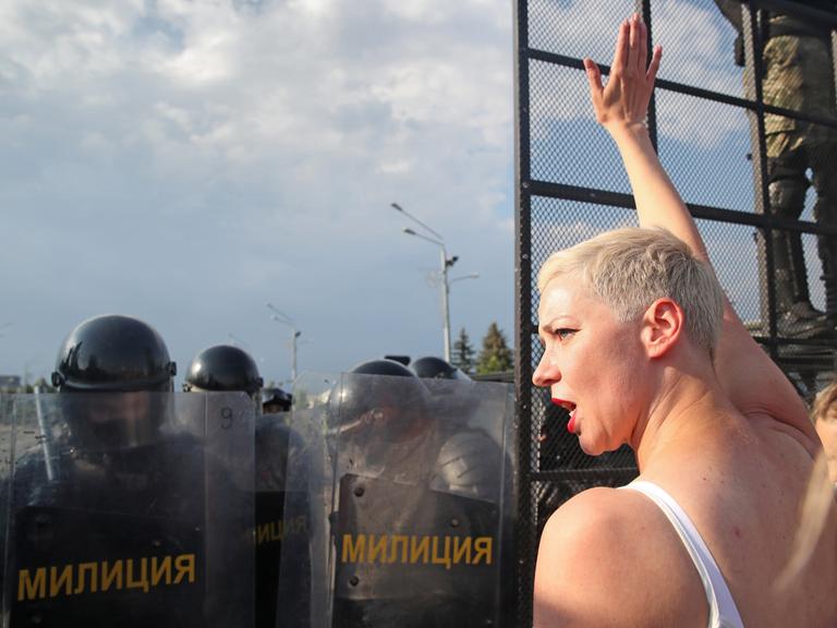 Die belarussische Bürgerrechtlerin Maria Kolesnikowa steht, bekleidet mit einem weißen ärmellosen T-Shirt, vor einer Riege von vermumten und bewaffneten belarussischen Sicherheitskräften. Sie recht die rechte Hand in die Höhe. 