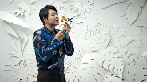 Ein Mann riecht mit geschlossenen Augen an einer Blumen. Er steht vor einer weißen Wand mit Papierblumen. 