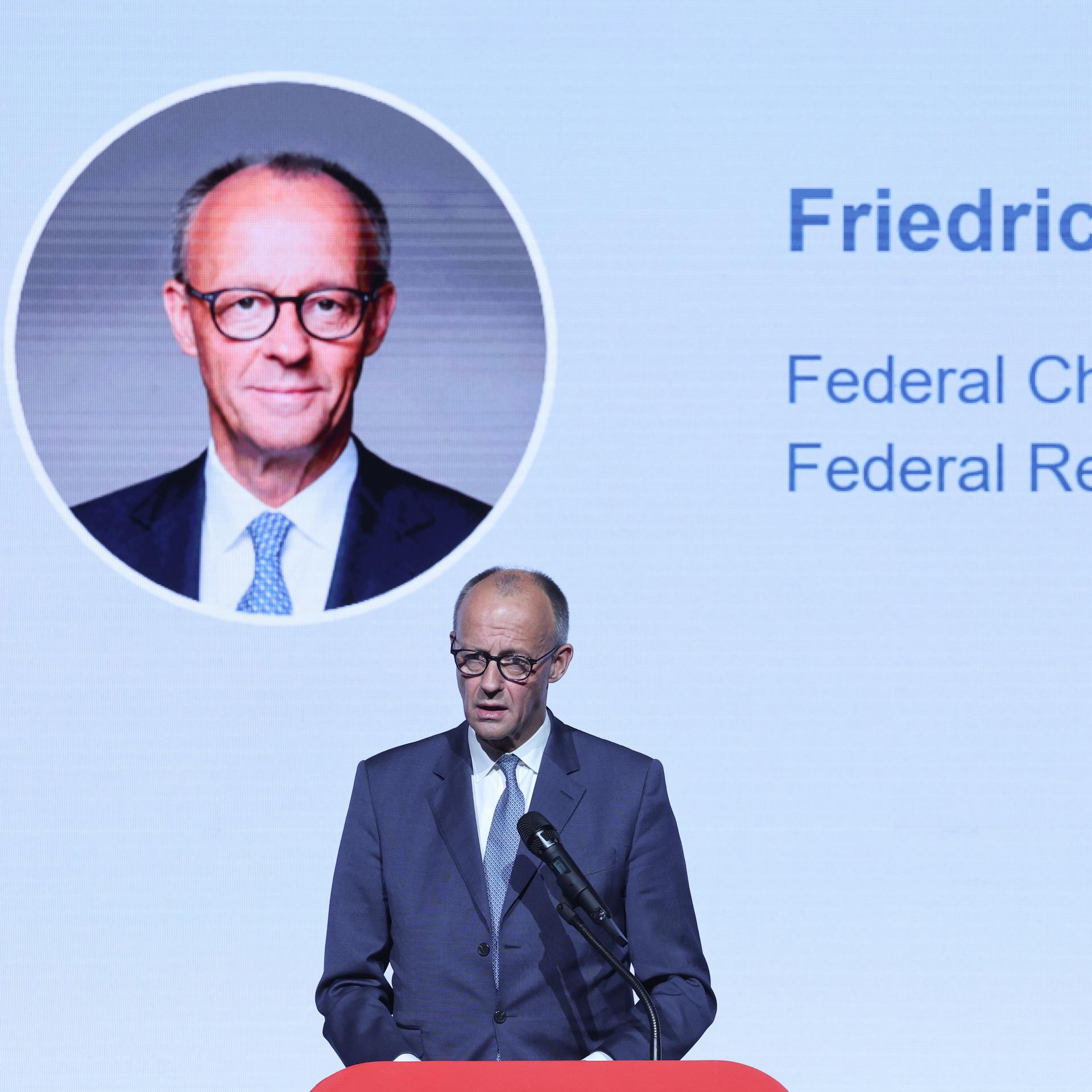 Zu sehen ist Bundeskanzler Friedrich Merz während einer Rede. Im Hintergrund ist ein großes Portaits von ihm an einer Wand zu sehen, daneben stehen auf englisch sein Name und seine Funktion zu lesen.