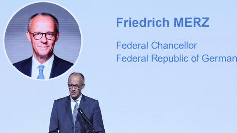 Zu sehen ist Bundeskanzler Friedrich Merz während einer Rede. Im Hintergrund ist ein großes Portaits von ihm an einer Wand zu sehen, daneben stehen auf englisch sein Name und seine Funktion zu lesen.