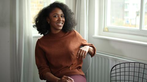 Natasha Brown sitzt in einem Braunen Strickpullover auf einem Stuhl und lächelt in die Kamera.