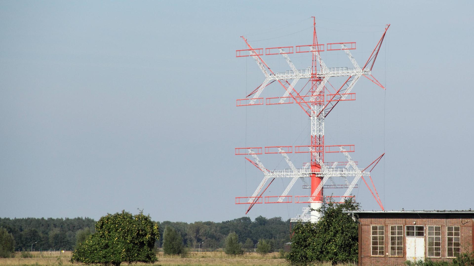 Das Bild zeigt eine in den Farben Rot und Weiß gehaltene große Kurzwellenantenne. Bezüglich der Bauart in Gitterstruktur erinnert das Gebilde an einen Strommast. Das Bild zeigt eine in den Farben Rot und Weiß gehaltene große Kurzwellenantenne. Bezüglich der Bauart in Gitterstruktur erinnert das Gebilde an einen Strommast.