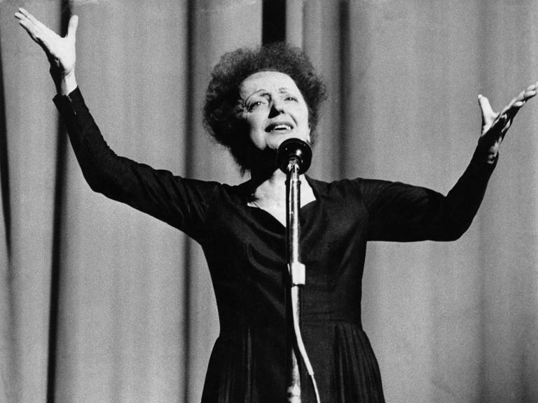 Edith Piaf steht bei einem Auftritt in Reims 1960 auf der Bühne vor einem Mikrofon und reckt die Arme in die Höhe.