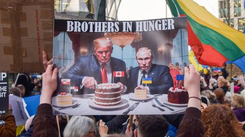 Ein Demonstrant vor der US-Botschaft in London hält im März 2025 ein Plakat hoch, auf dem Trump und Putin Kanada- und Ukraine-Kuchen essen.