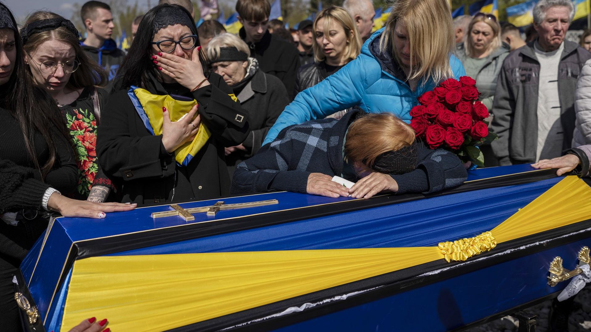 Trauernde Frauen drängen sich um einen Sarg, der in den Nationalfarben der Ukraine, Blau und Gelb, geschmückt ist, weitere Frauen und Männer stehen im Hintergrund.