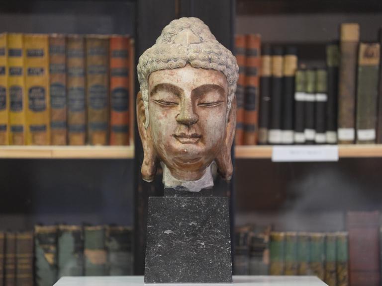 Ein Buddha-Kopf aus Marmor steht in einem Raum des Hamburger MARKK-Museums am Rothenbaum. Er steht auf einem dunklen Marmorklotz. Im Hiintergrund stehen historische Bücher im Regal. 