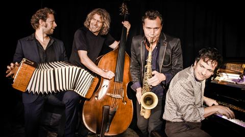 Vier Musiker spielen vor einem schwarzen Hintergrund. Die Männer musizieren auf Bandoneon, Kontrabass, Saxophon und Klavier. Nur der Saxophonist blickt direkt in die Kamera, die anderen schauen sich gegenseitig an oder zur Seite.
