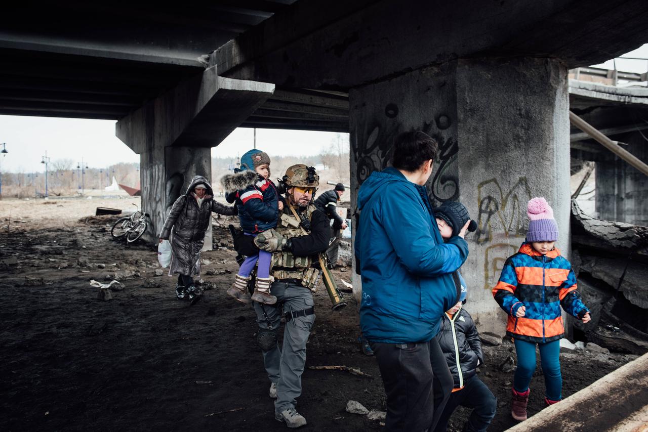 Ein Soldat trägt ein Kleinkind auf dem Arm, der Vater des Kleinkinds wartet mit einem Baby auf dem Arm, daneben ein weiteres Kind. Die Gruppe steht unter einer Brücke. Es sind Zerstörungen zu sehen.