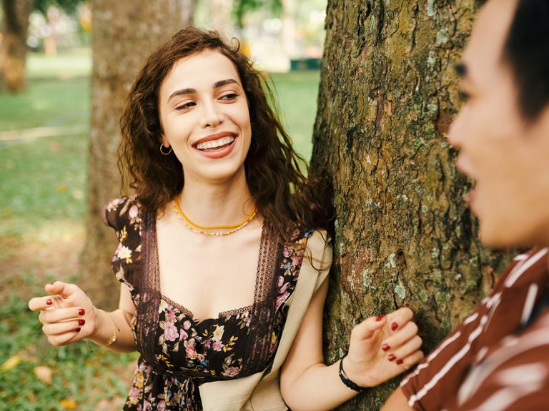 Eine junge Frau lehnt an eine Baum im Park, neben ihr ein junger Mann. Man könnte es als Flirt interpretieren.