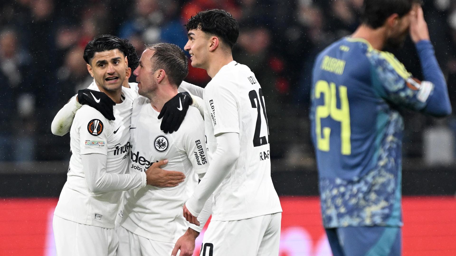 Die drei Spieler in den weißen Trikots und in weißen Hosen umarmen sich. Daneben ein enttäuschert Ajax-Spieler im gelb-blauen-Trikot, der sich mit der Hand die Stirn abwischt. Im Hintergrund unscharf eine Zuschauertribüne. 