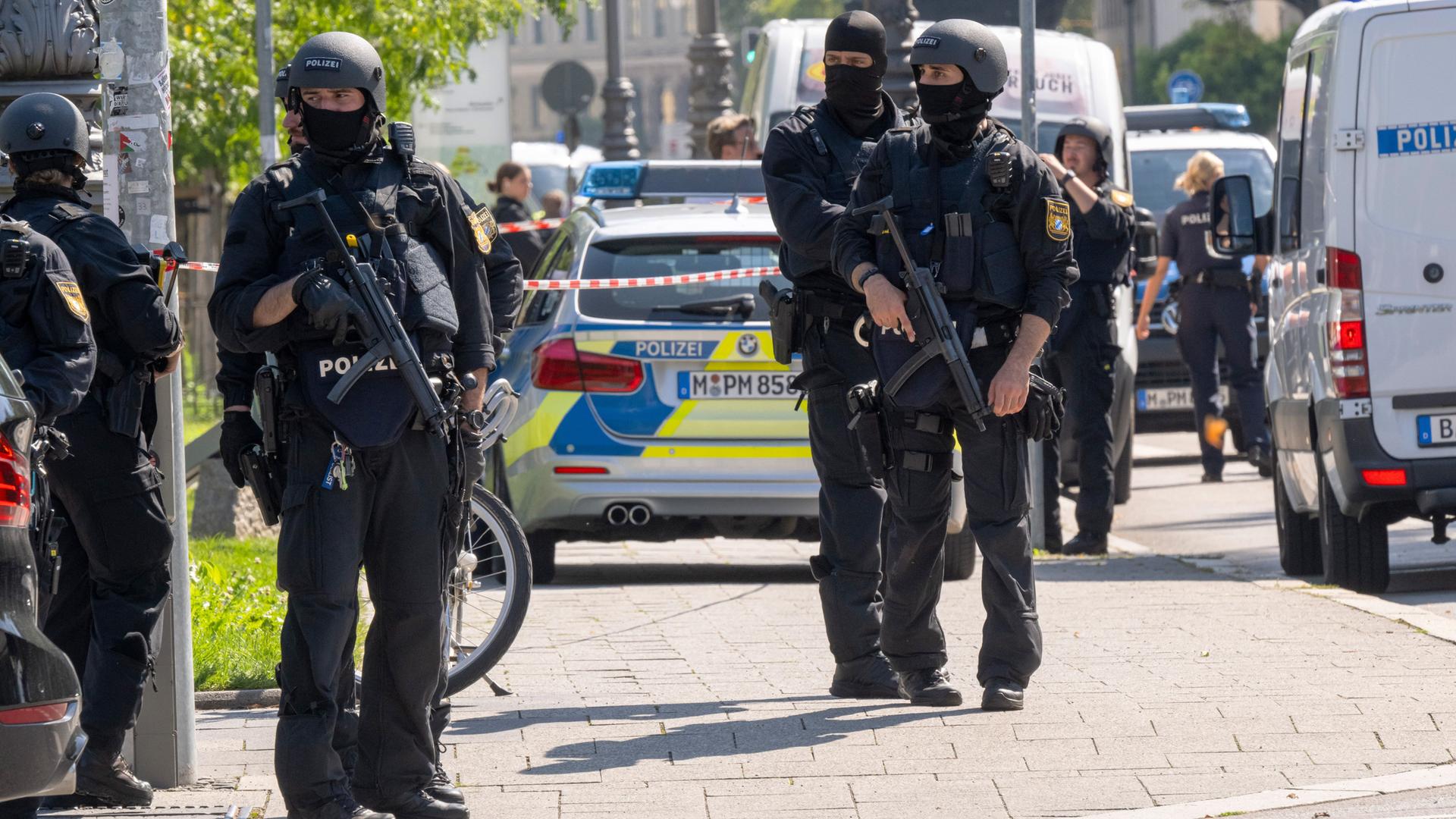 Polizisten im Einsatz in München. Die Polizisten sind schwer bewaffnet und vermummt. Sie stehen auf einem Bürgersteig vor einem Polizeiwagen.