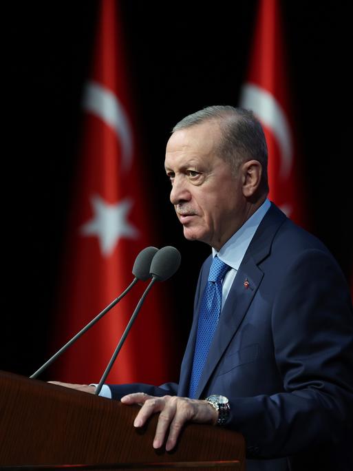 Recep Tayyip Erdoğan steht im blauen Anzug an einem Rednerpult und spricht. Im Hintergrund sind mehrere türkische Flaggen zu erkennen.