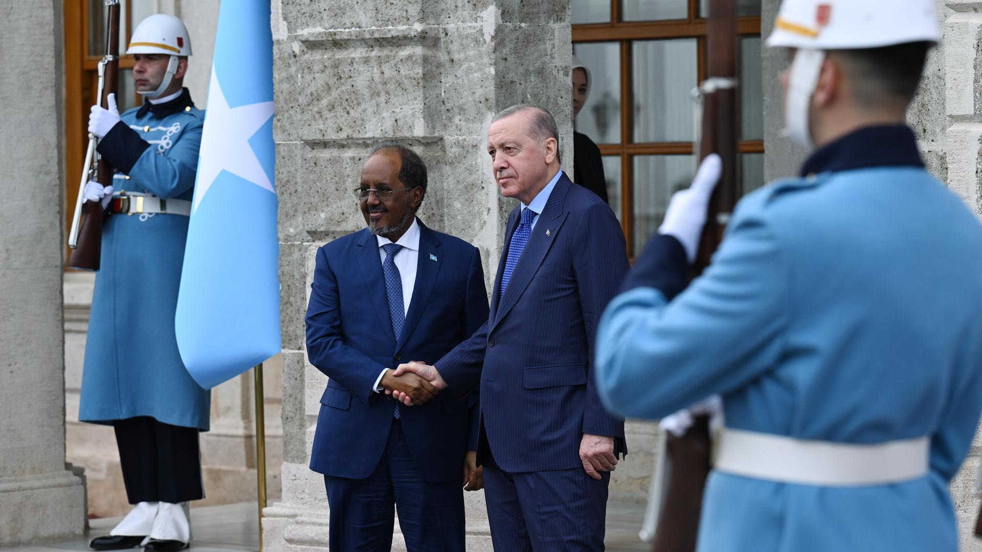 Recep Tayyip Erdoğan und Hassan Sheikh Mohamud schütteln sich bei einem offiziellen Termin die Hände. Die Szene ist mit der blau-weissen Flagge Somalias und Soldaten in Uniform gerahmt.