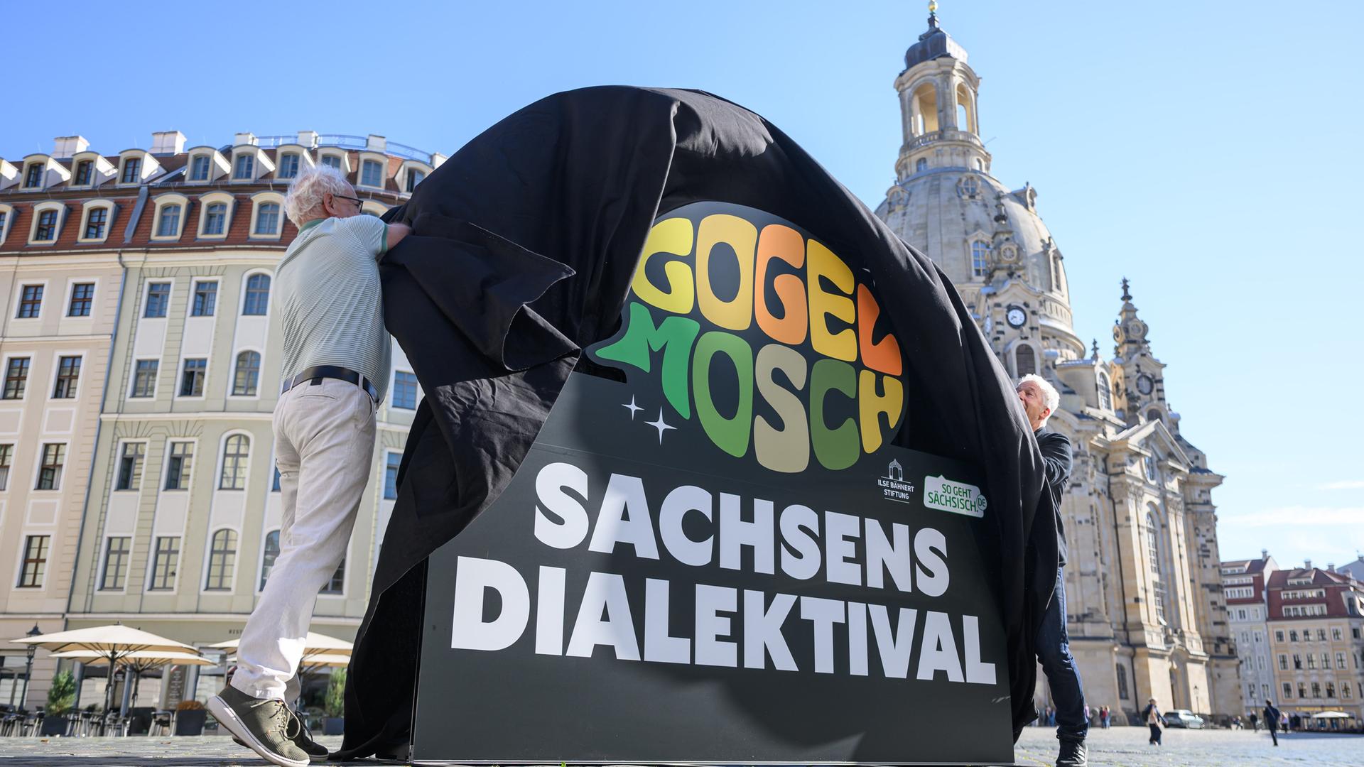 Tom Pauls (l), Schauspieler und Kabarettist, und Autor Peter Ufer enthüllen bei einem Pressetermin zur Vorstellung des Dialektfestivals "Gogelmosch" auf dem Neumarkt vor der Frauenkirche einen Aufsteller mit dem Logo der Veranstaltungsreihe. 