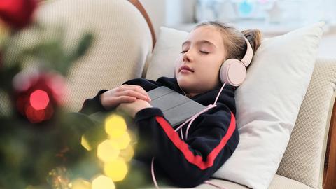 Ein Mädchen liegt entspannt mit Tablet und Kopfhörern auf dem Sofa. Im Vordergrund ist unscharf ein geschmückter Weihnachtsbaum zu sehen.