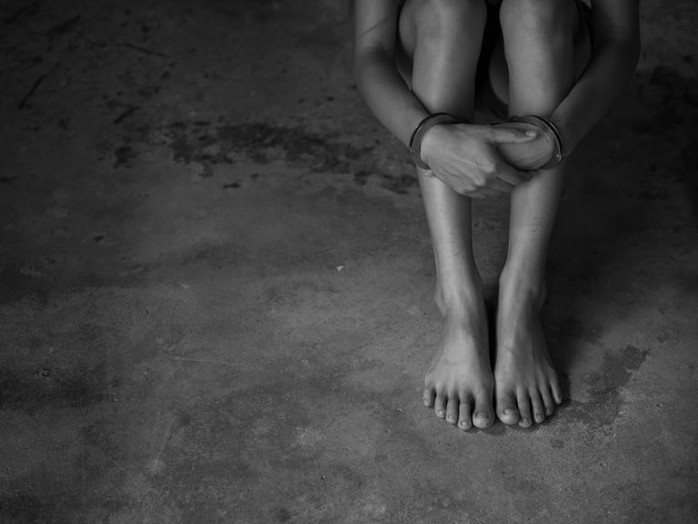 Eine Person sitzt mit Handschellen auf dem Boden. Zu sehen sind nur Arme und Beine. Das Foto ist schwarz-weiss.