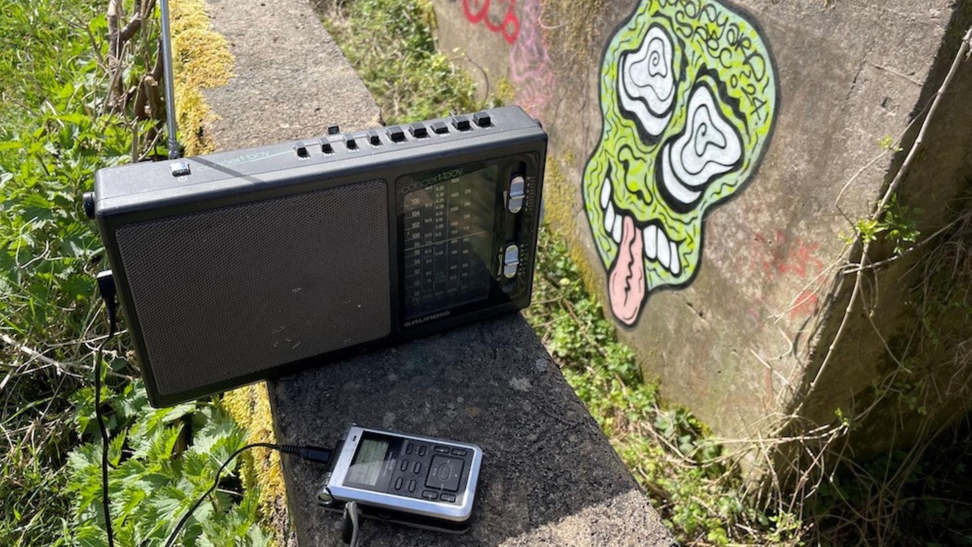 Ein altes Radio und ein Aufnahmegerät in der Natur, auf einer Steinwand im Hintergrund ein Graffito. Ein altes Radio und ein Aufnahmegerät in der Natur, auf einer Steinwand im Hintergrund ein Graffito.