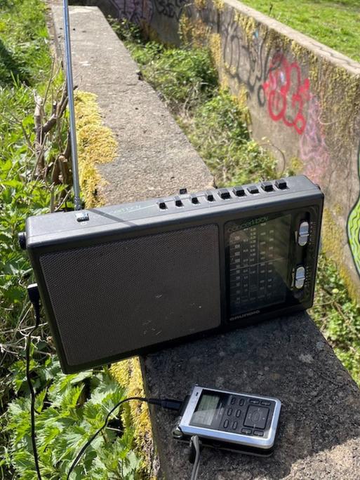 Ein altes Radio und ein Aufnahmegerät in der Natur, auf einer Steinwand im Hintergrund ein Graffito.