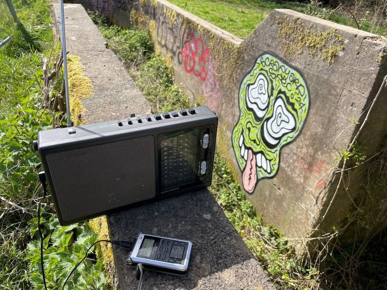 Ein altes Radio und ein Aufnahmegerät in der Natur, auf einer Steinwand im Hintergrund ein Graffito.