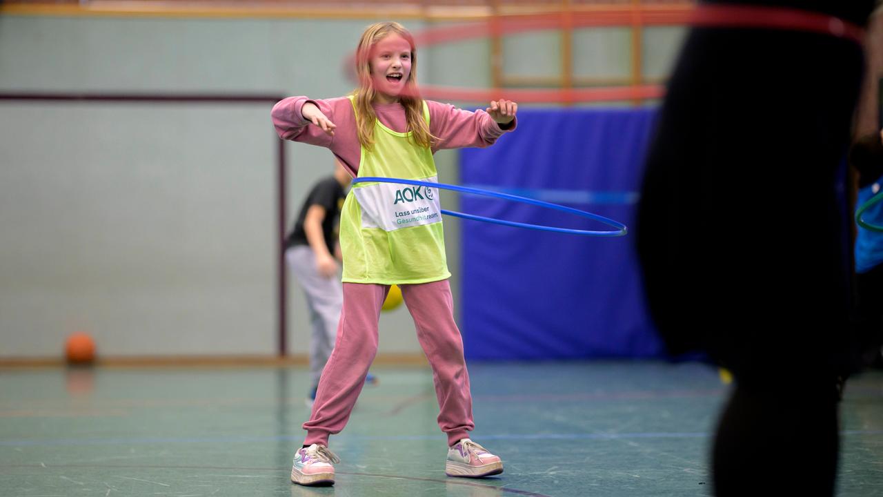 Ein blondes Mädchen im Grundschulalter steht in einer Turnhalle, trägt ein gelbes Leibchen über dem rosafarbenen Sportanzug und schwingt einen Hula-Hoop-Reifen um ihren Bauch. 