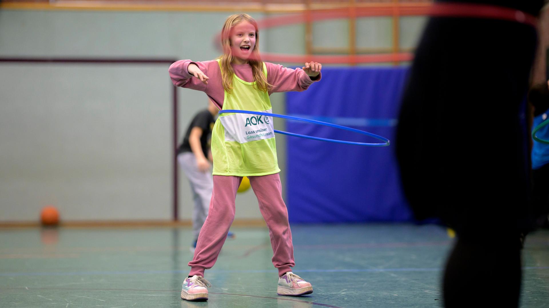 Ein blondes Mädchen im Grundschulalter steht in einer Turnhalle, trägt ein gelbes Leibchen über dem rosafarbenen Sportanzug und schwingt einen Hula-Hoop-Reifen um ihren Bauch. 
