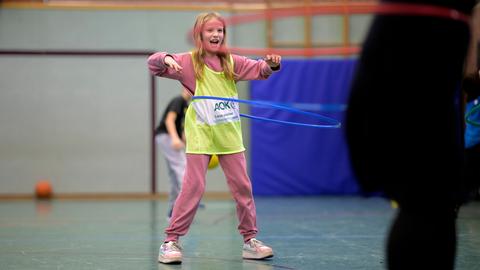 Ein blondes Mädchen im Grundschulalter steht in einer Turnhalle, trägt ein gelbes Leibchen über dem rosafarbenen Sportanzug und schwingt einen Hula-Hoop-Reifen um ihren Bauch. 